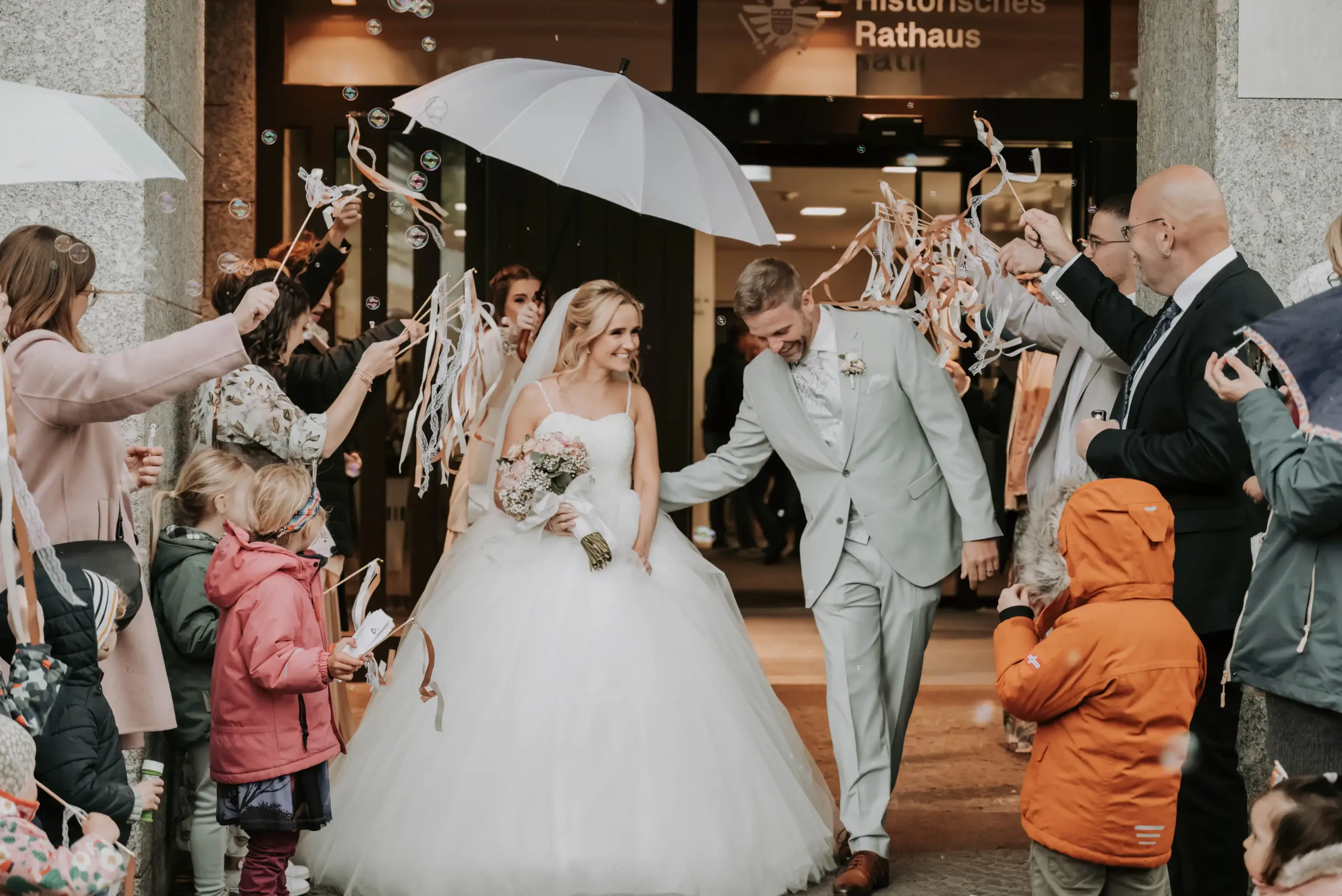 Selina und Marc Philip laufen nach der Trauung durch jubelnde Gäste, während eine Trauzeugin einen Schirm über ihnen hält