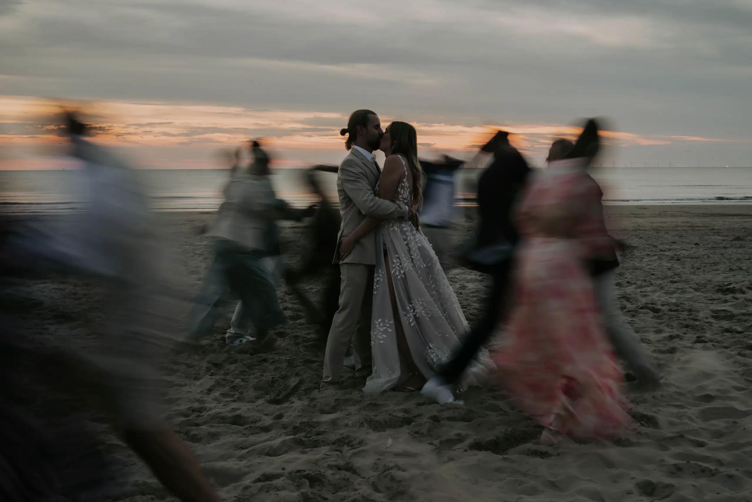 Brautpaar steht still am Strand, während Gäste durch eine Langzeitbelichtung verschwommen durchs Bild laufen